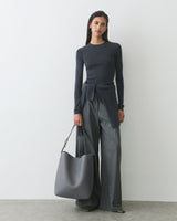 A person stands holding a large shoulder bag, wearing a long-sleeve top and wide-leg trousers.