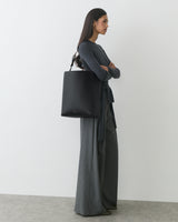 Person in long-sleeve top and wide-leg pants holding large tote bag standing against white wall
