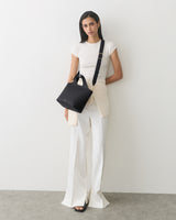 A person standing with a shoulder bag, wearing a short-sleeve shirt and wide-leg pants.