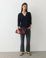 Person holding a handbag, wearing a sweater, jeans, and sandals standing in front of a plain wall.