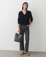 Person wearing sweater and jeans, holding a handbag, standing against plain background.
