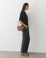 Person standing in profile with a small shoulder bag, hands in pockets, wearing casual attire.