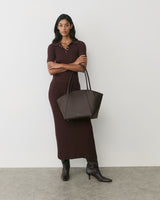 Person in long dress holds large handbag, wearing boots against plain backdrop.
