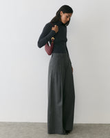 Person standing and looking down, holding a small shoulder bag, wearing wide-leg pants.