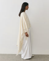 Person wearing cape and long skirt stands in profile against plain background.