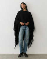 Person wearing a cape with fringe detail and jeans, standing against a plain background.