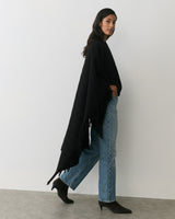 Person wearing cape, jeans, and ankle boots standing against wall.