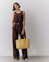 Person standing with tote bag and hands in pockets against plain background.