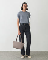 Person in a shirt and jeans holding a handbag standing on a plain background.