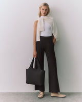 Person standing, wearing a top, pants, with a sweater over shoulders, holding a tote bag.