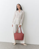 A person stands holding a large handbag, wearing a long-sleeve shirt and wide-leg pants.