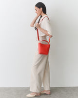 Person standing, holding a red shoulder bag, wearing a shirt and wide-leg pants.