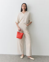 Person in relaxed outfit holding a small bag while leaning against a wall.