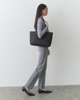 Person standing with a tote bag on shoulder, wearing a long-sleeve top and pants.