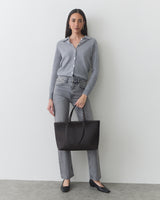 Person in casual outfit holding a tote bag standing against a plain backdrop.