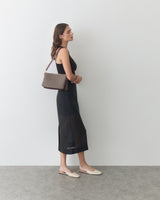 Woman in dress holding a shoulder bag, standing sideways against a plain wall.
