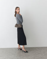 Woman in long skirt with shoulder bag and heels stands against plain wall.