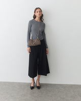 Woman stands against wall wearing long-sleeve top, wide-leg pants, crossbody bag, and heels.