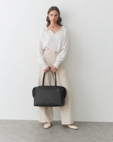 Person standing, holding a handbag, wearing a long-sleeve top and wide-leg trousers.