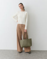 Person stands with large tote bag, wearing a sweater and wide-leg pants.