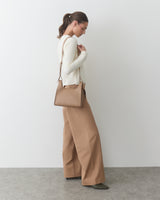 Person standing, holding a shoulder bag, wearing a long-sleeve top and wide-leg pants.