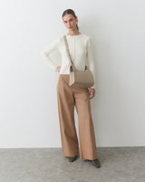 Person standing against wall holding a handbag, wearing a long-sleeve top and wide-leg pants.