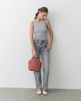 Person in tank top and jeans holding a handbag, standing against a plain backdrop.