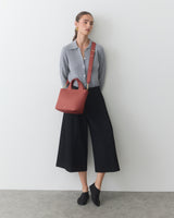Person wearing a cardigan, culottes, holding a shoulder bag, leaning against a wall.