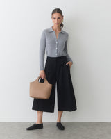 Person holding a handbag, wearing a long-sleeve top and wide-leg pants, standing indoors.