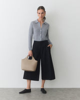 Person posing with wide-leg pants, buttoned top, holding a handbag, hands in pockets.