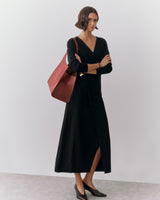 Person in long dress with shoulder bag and heels, standing against plain backdrop.