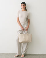 Person in simple outfit holding a tote bag, standing against a plain wall.