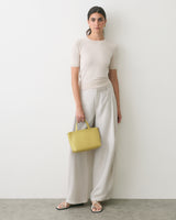 Person standing against wall holding a handbag wearing a short-sleeved top and wide-legged pants.