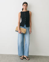Woman in sleeveless top and jeans holds woven clutch, wearing sandals.