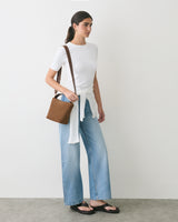 Person wearing a t-shirt, jeans, sandals, with a shoulder bag, standing against a plain wall.