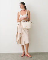 Person in a dress holding a handbag, wearing sandals and a necklace, standing against a plain wall.
