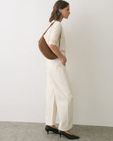 Woman in light outfit poses sideways with shoulder bag and heels.