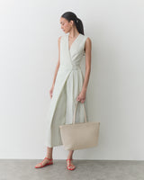Person in sleeveless dress holding a large handbag, wearing sandals, standing on a plain background.