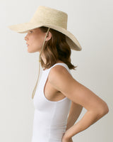 Person in a sleeveless top wearing a wide-brimmed hat, facing left, hands on hips.