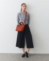 Person in long pants and shirt, carrying a shoulder bag, standing against light background.