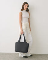 Person standing against a wall holding a large handbag, wearing a sleeveless top and long pants.