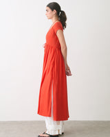 Woman in a long dress over pants, standing sideways in a neutral room.