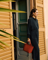 Person in a relaxed jacket and pants holding a handbag, standing by an open door.