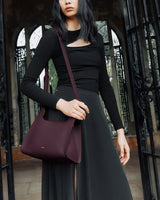 A person holding a shoulder bag, standing in front of arched windows.