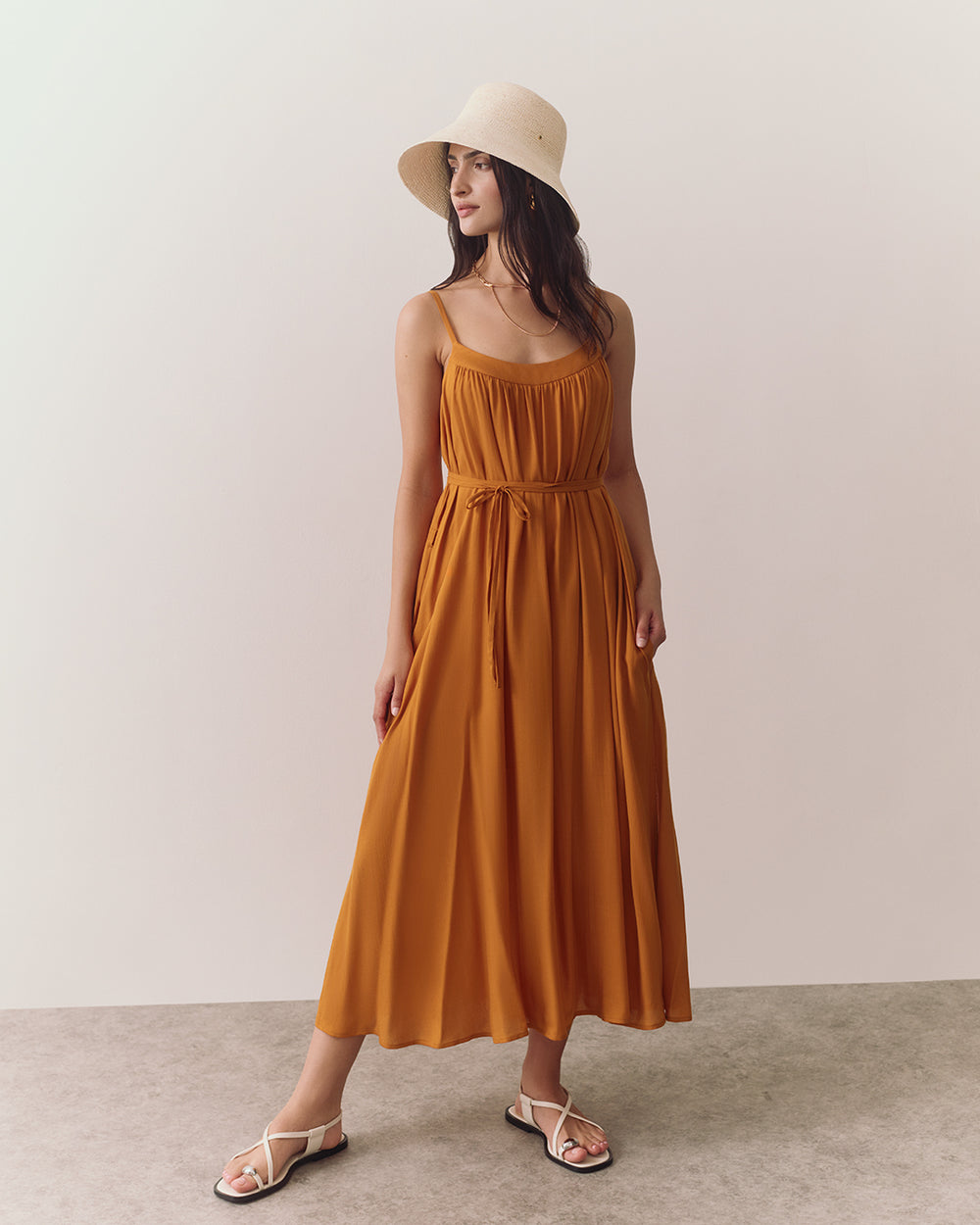 Woman in a long dress and wide-brim hat stands on a plain background.