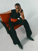 Woman sitting on chair leaning head on hand, wearing pants and top with boots.