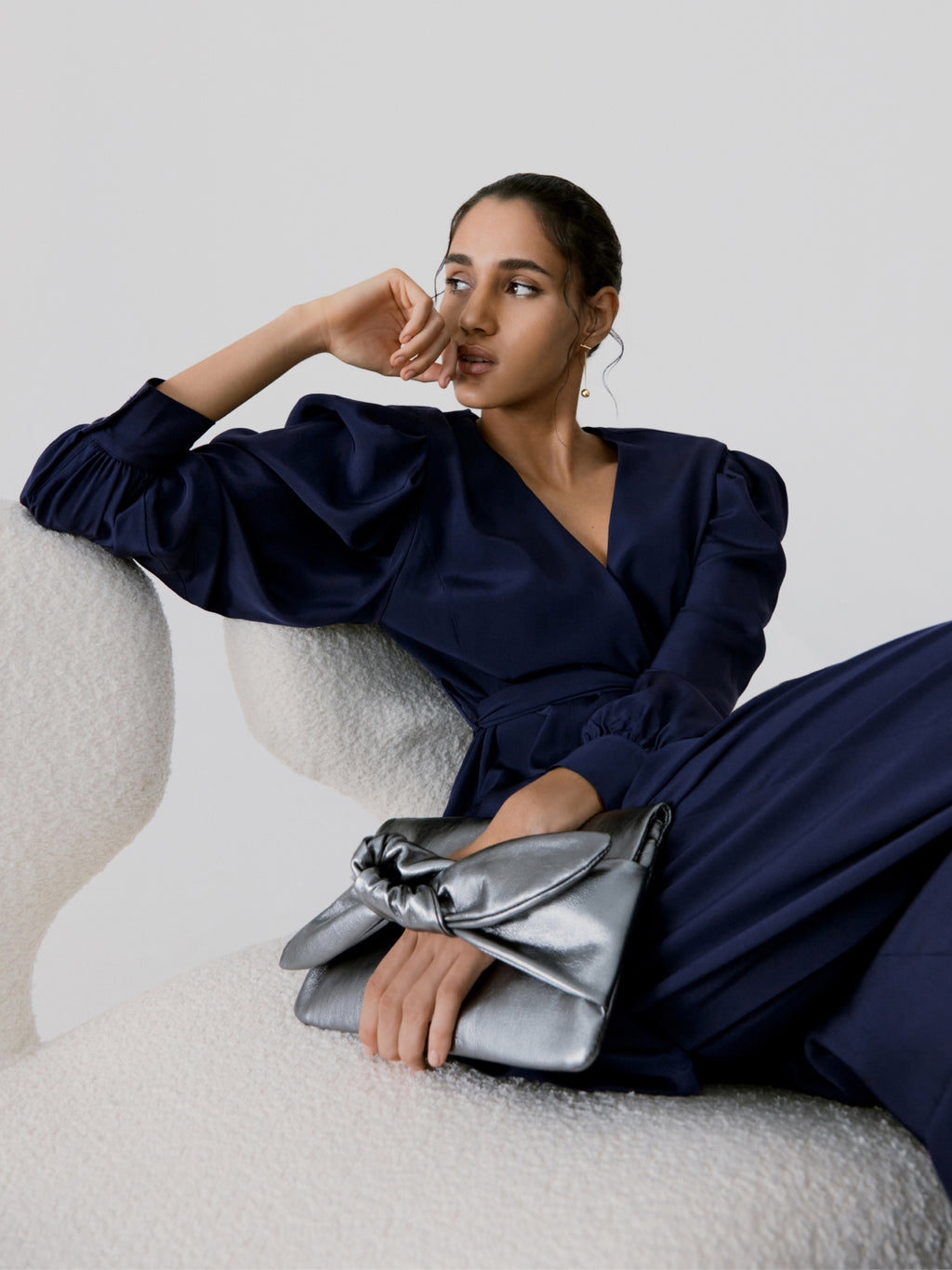 Woman reclining on curved chair with hand on cheek and holding a clutch.