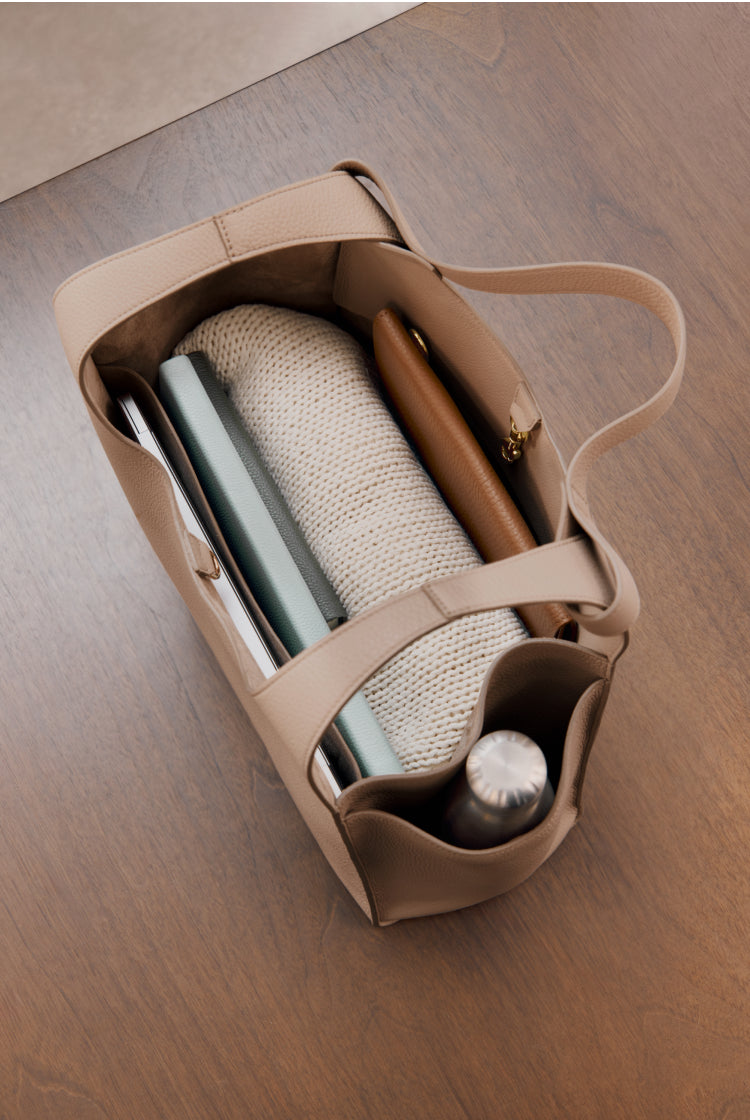 Interior of a large beige tote with a laptop, notebooks, a sweater, a water bottle and a wallet placed inside. 
