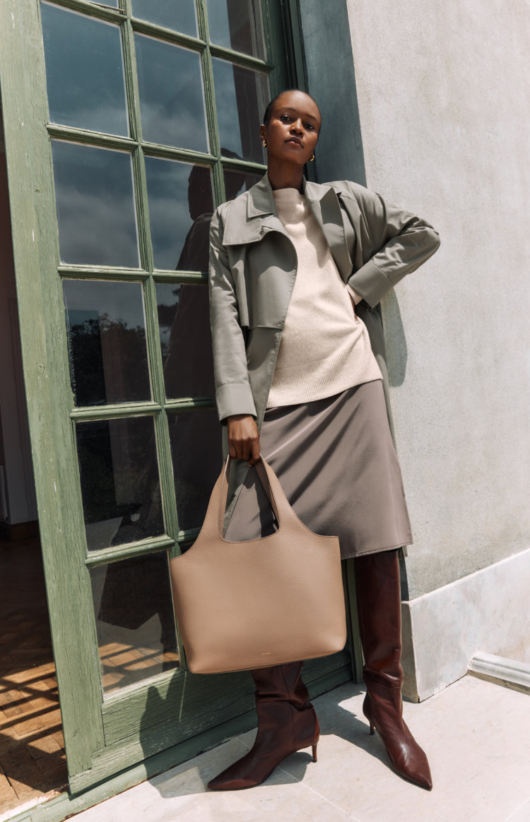 Woman standing by a window holding a large bag, wearing a jacket, skirt, and boots.
