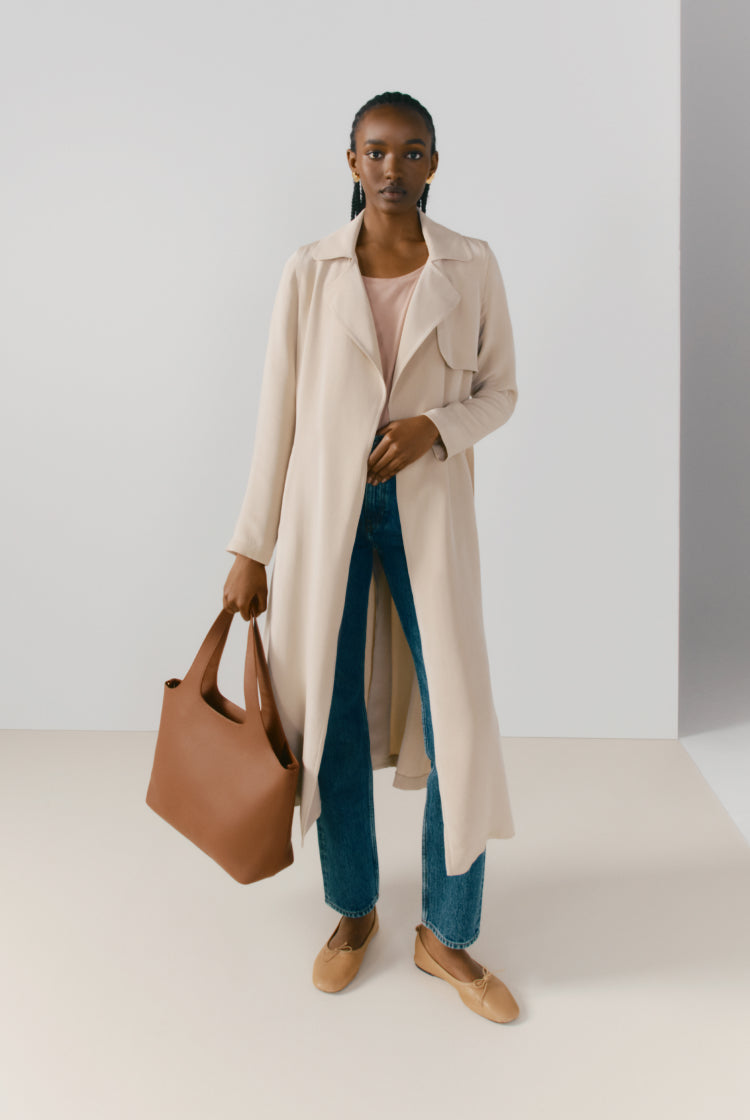 Woman standing in a studio holding a bag and wearing a long coat and jeans.